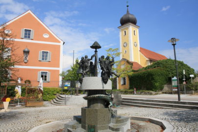 Dorfbrunnen am Kirchplatz in Wald mit Kirche und Gebäude im Hintergrund.