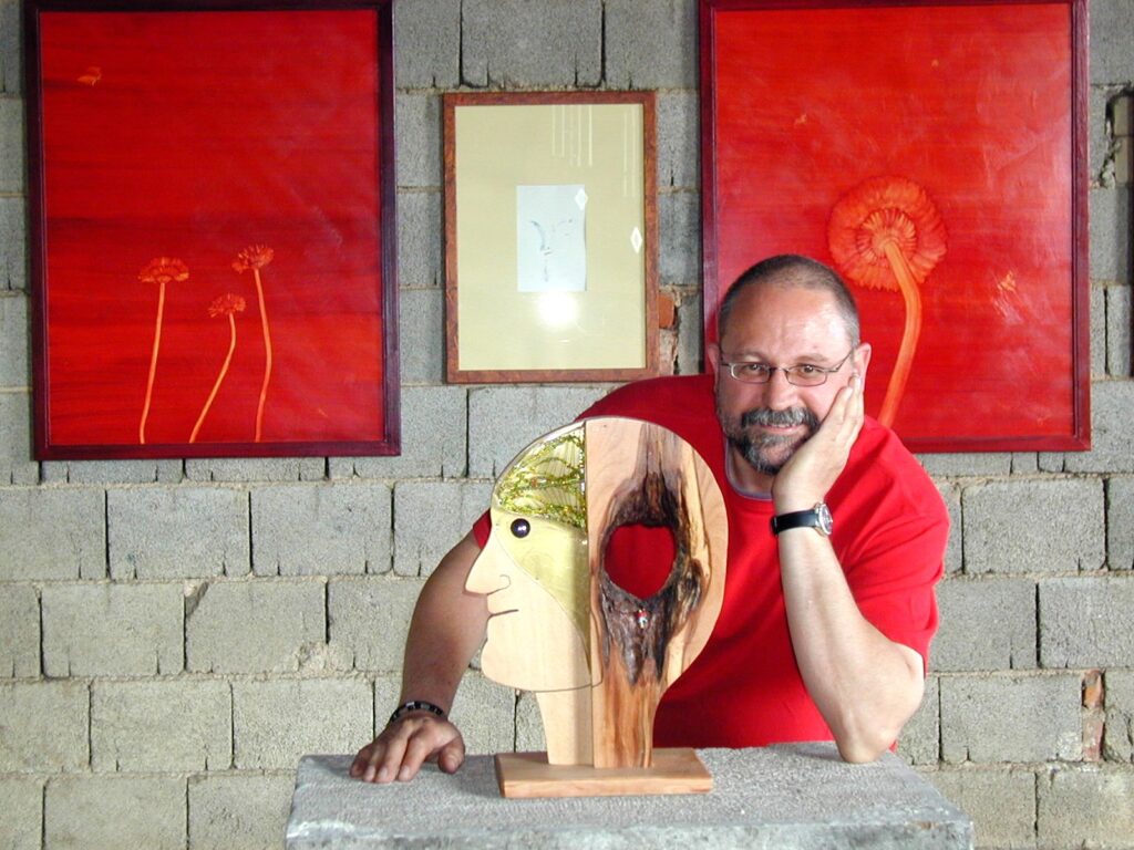 Mann, namens Sam Stadler, im Hintergrund drei Bilder mit Blumen und im Vordergrund eine Skulptur, die einen Kopf aus Holz zeigt.