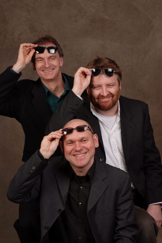  Drei Männer mit Anzung und hochgeklappter Sonnenbrille bilden die Musikgruppe Trio Salato. 